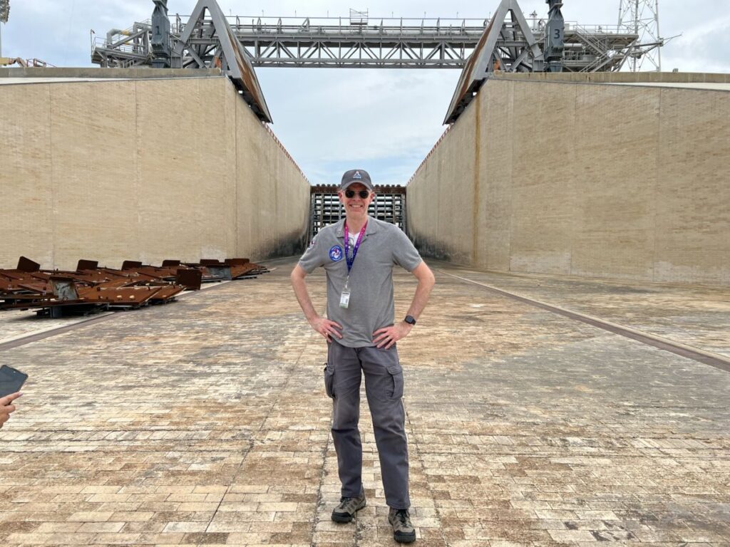View Larger Image (The author standing in the flame-trench of Pad 39-B at the Kennedy Space Center. Directly behind me is the flame-deflector which diverts the exhaust from the SLS rocket’s four RS-25 engines and two solid-rocket boosters, equally to each side of the deflector. In the foreground on the left side of the image, stacked on the ground, are the steel panels that have been removed from the flame-deflector. These were damaged by the greater than anticipated energy from the November 2022 launch of Artemis 1. These will all be replaced with new and stronger panels before the launch of Artemis II, currently scheduled for the end of 2023)
