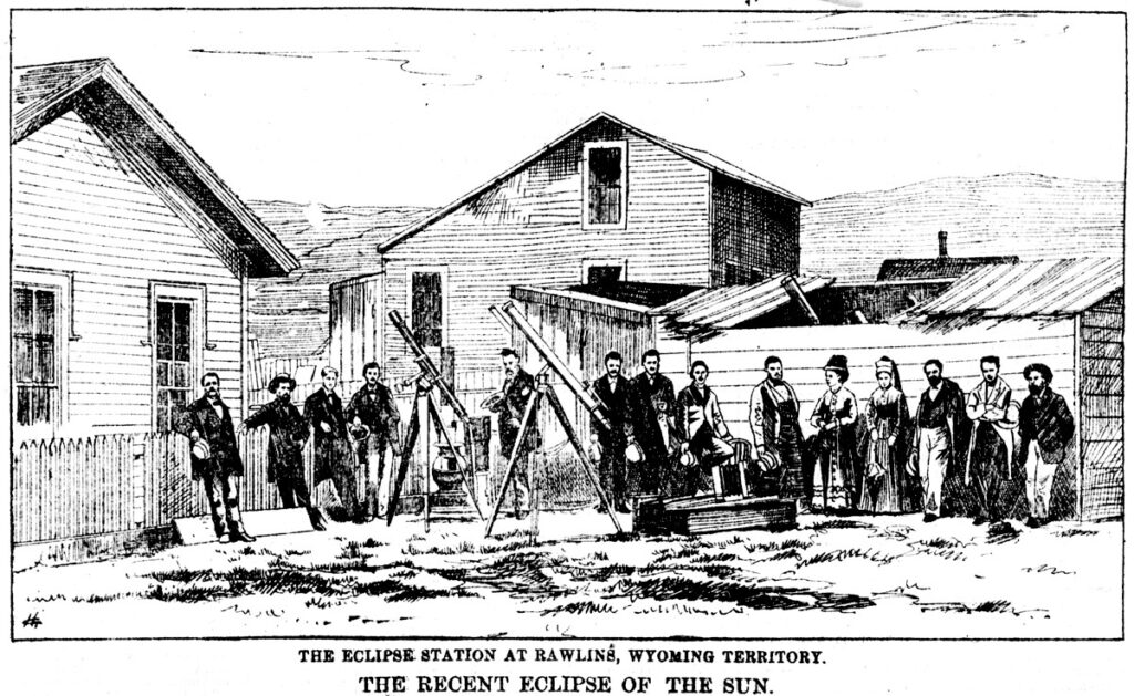 A dozen men and two women stand along a fence or lean against a shed. Two telescopes sit in the foreground.