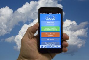 Cloud App on phone being held by a hand up to the sky. Clouds in the sky in the background. 